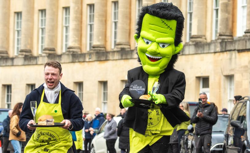 A man dressed in Regency costume and a man dressed in a Frankenstein costume running along Bath's Royal Crescent while holding trays containing a champagne flute and bun.