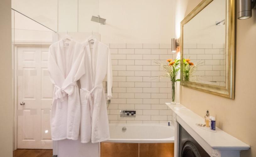 A white and gold bathroom with two bathrobes hanging on the shower - Credit Evoke Pictures