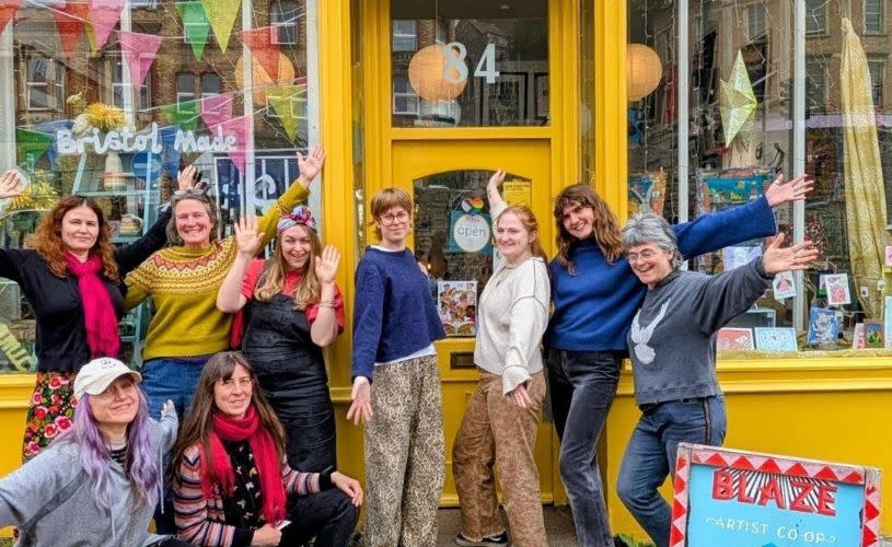 Blaze Independent Shop Front. Yellow shop with colourful objects in the window and 9 nine people stood out the front of the shop wearing colourful clothes and all smiling with their arms held out wide.