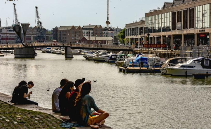 People sitting on the Bristol Harbourside with Pero's Bridge and the cranes outside M Shed in the background