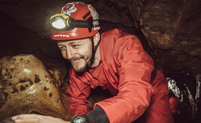 A man wearing a red caving helmet and overalls places his hand on a rock as he moves through a cave in the Mendip Hills. He is smiling and looks relaxed.