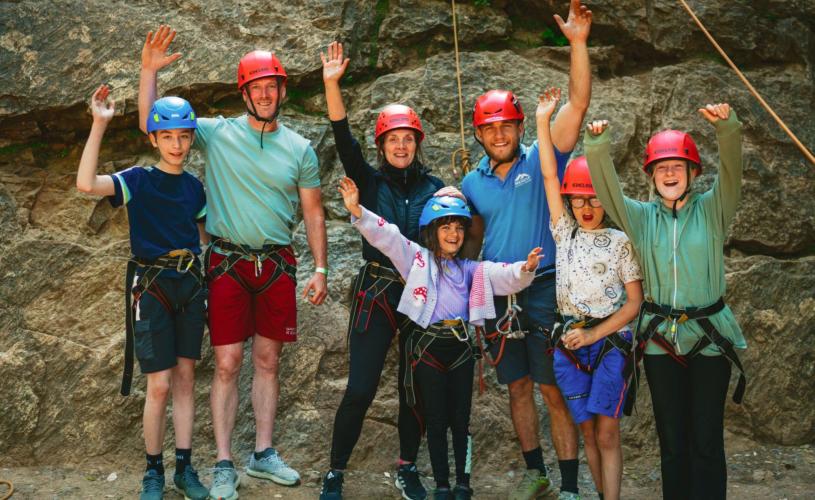 A family of two adults and four children stand in a group with their rock climbing instructor at the bottom of a rockface. They are wearing red helmets and black climbing harnesses. They are raising their arms and smiling, looking proud and exhilarated by their rock climbing activity.