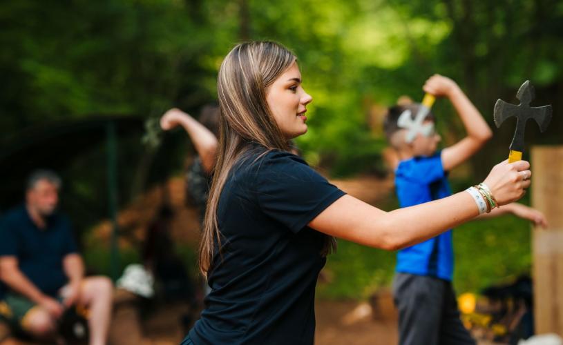 A teenager is aiming her axe at a target during an axe-throwing target sports activity at Mendip Activity Centre. She is wearing a black shirt and has a look of concentration on her face.