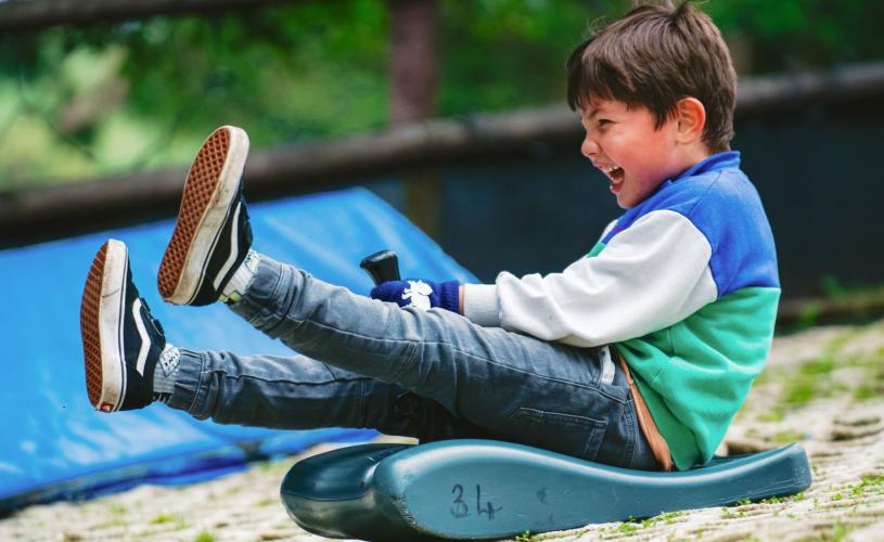 A boy on a blue toboggan kicks his feet up, smiling and laughing as he slides down the all-weather toboggan slope at Mendip Activity Centre.