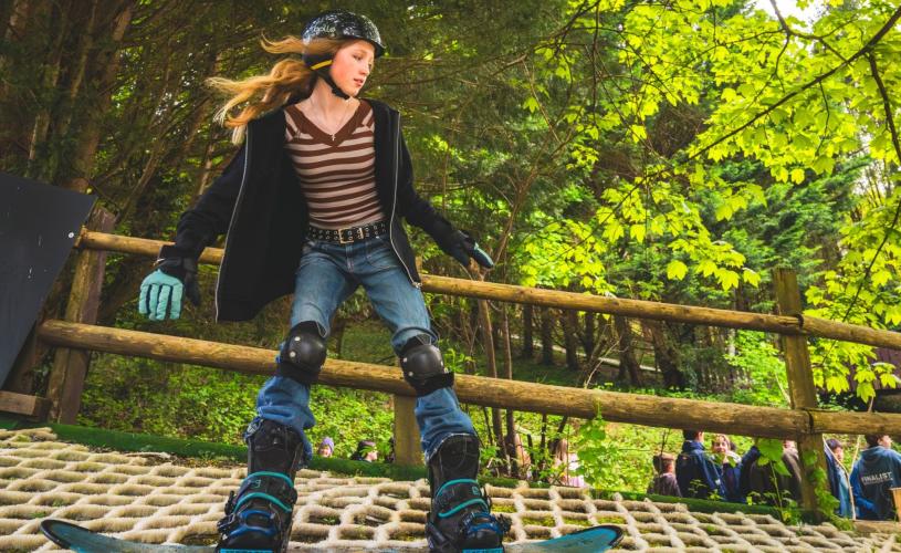 A young person rides a snowboard down Somerset’s only dry slope. Her arms are slightly raised to her sides for balance, and she is wearing a helmet, gloves and knee pads. Her hair streams out behind her in the wind as she carves down the slope.