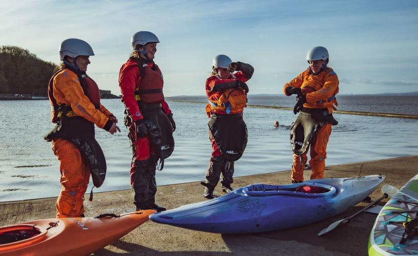 Four women stand in a row next to kayaks and paddleboards, taking a break while on an outdoor activity course. They are talking while standing on the edge of a marine lake.