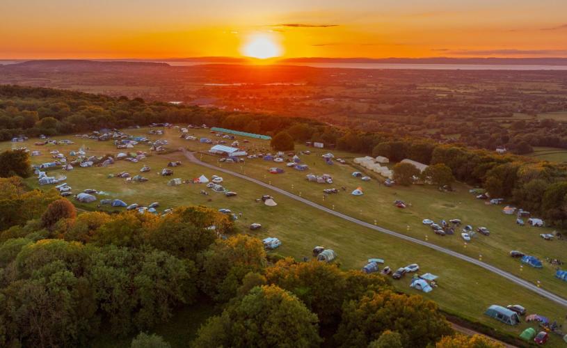 Aerial view of Mendip Basecamp at sunset, looking peaceful and serene. The large family campsite field is circled by woodland, with several tents and campervans dotted around.