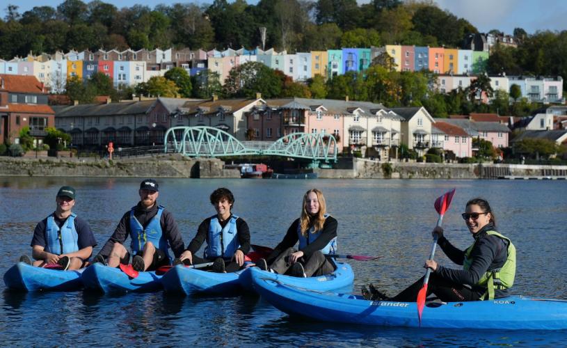 Five people in sit-on-top kayaks on Bristol Harbour pose together for a photo with Bristol’s famous colourful terrace houses on the hill in the background. They are smiling and look relaxed, having a good time on a unique urban adventure.