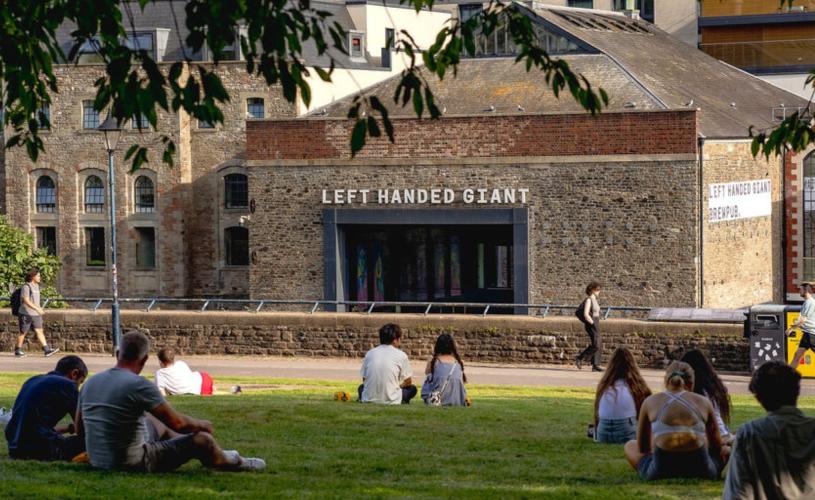 People sitting on the grass in Castle Park with the Left Handed Giant Brewpub on the opposite side of the river in the background