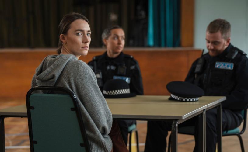 A woman in a grey hoodie sits at a table in a large hall, looking towards the camera. Two uniformed police officers sit across the table, with two police hats placed in front of them.