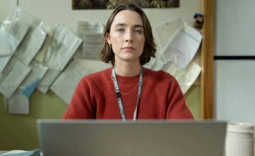 A woman wearing a red jumper and staff lanyard sits behind a laptop at a desk, looking towards the camera. Papers are pinned to the wall behind her and a takeaway cup sits on the desk.