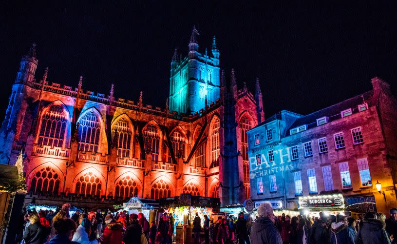 Bath Abbey lit up in rainbow colours with Bath Christmas Market in the foreground
