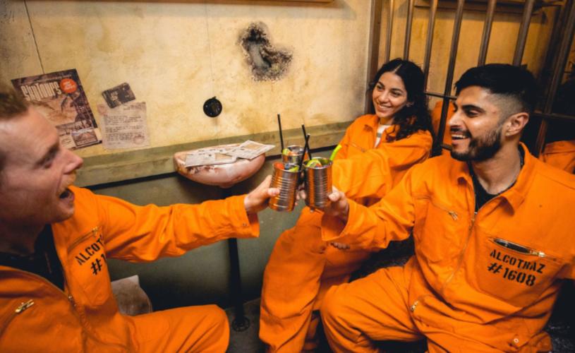 Group with cocktails wearing orange jumpsuits in a cell - credit Alcotraz