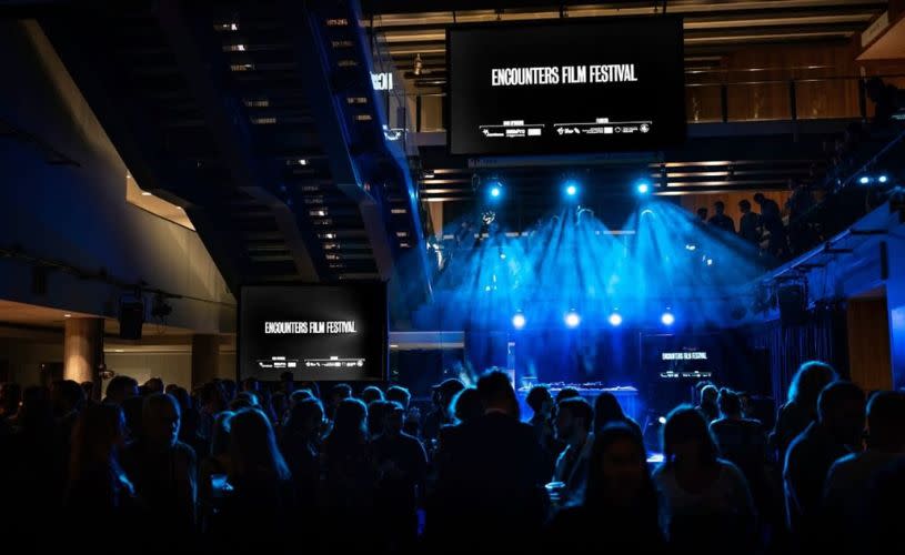 A group of people in a dark room with blue lights and a screen that says Encounters Film Festival