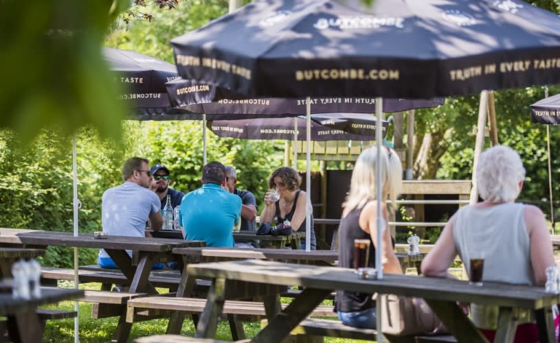 People sat on wooden benches with sun umbrellas that say Butcombe on the side - Credit Butcombe Brewery