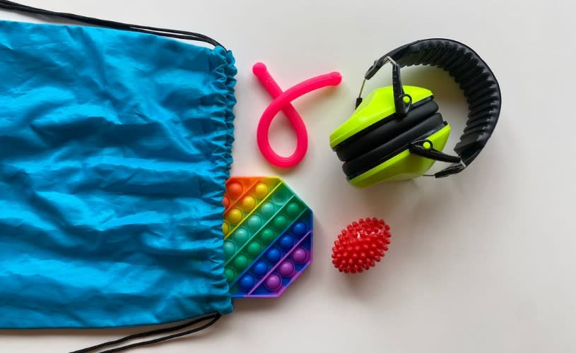 A blue string backpack with a rainbow pop it, a pink tube, a red spiky ball and a pair of yellow ear defenders