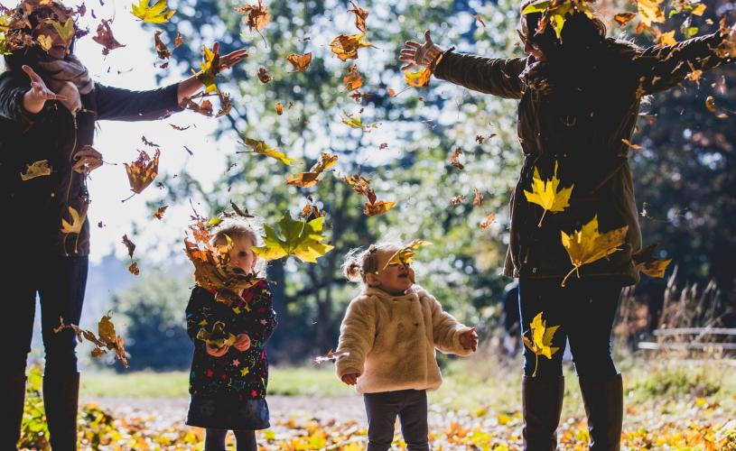 Two mothers and their young daughters throwing fallen leaves in the grounds of Westonbirt Arboretum near Bristol in autumn - credit Johnny Hathaway