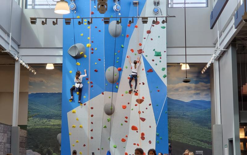 Two people climbing a blue and gray rock wall while a group of people watch at the bottom