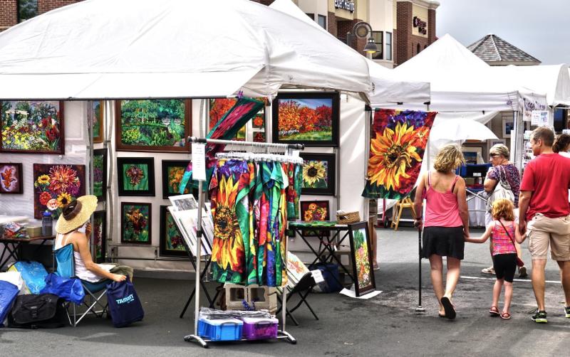 A family walks past a white tent filled with colorful t-shirts and paintings at Arbor Lakes Art Fair