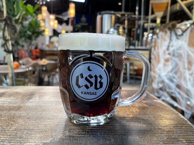 Small beer glass beer mug filled with dark beer. The mug has a logo that reads "CSB Kansas"