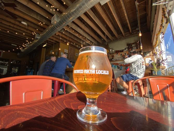 Full beer glass with the words "Brewed for the Locals" sits on a table with the bar in the background at Hopping Gnome Brewing Company