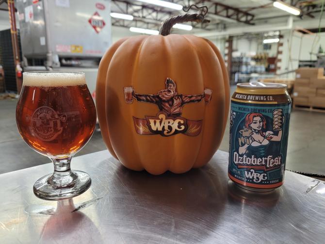 A pumpkin with a drawing of a man holding two beers and the letters WBC sits on a table next to a glass of beer and a can of beer that says "Oztoberfest"