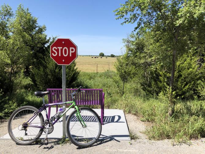 A green bike rests against a stop sign with a purple park bench behind it overlooking a farm scene