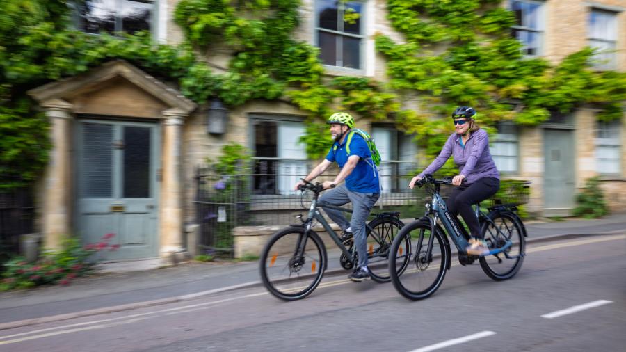Two cyclists riding bikes through picturesque Charlbury, passing stone cottages covered with greenery