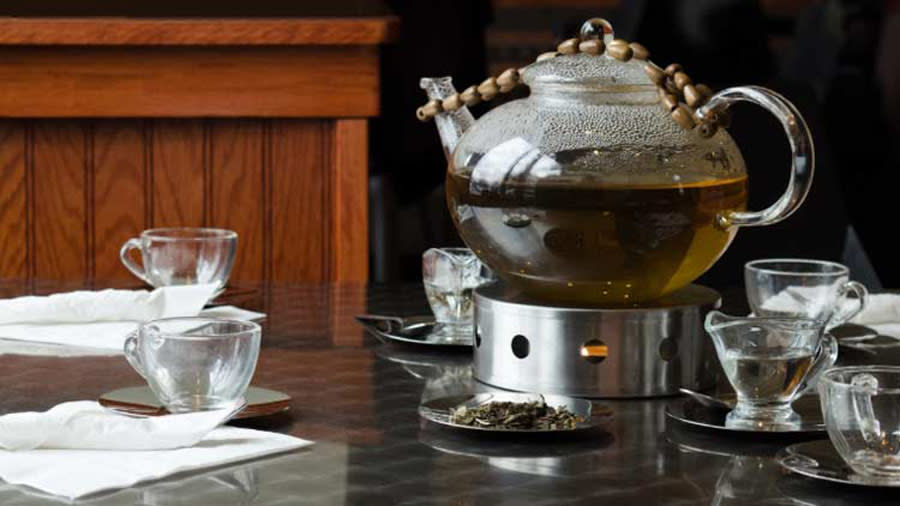 A grand teapot filled with black tea, and six teacups sitting around it on an oak table