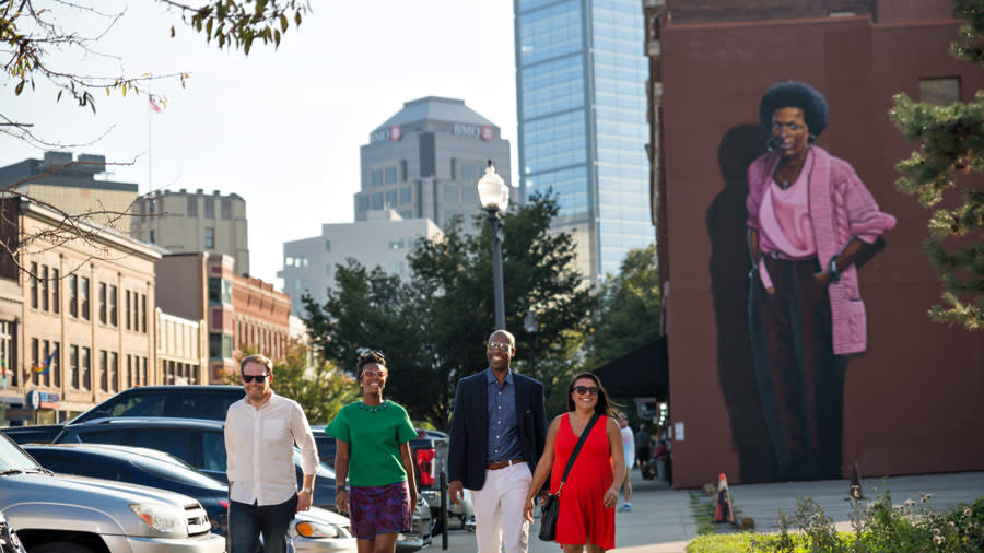  A group of people walking, smiling on the sidewalk. Behind them, a larger-than-life mural of poet Mari Evans looks over the street
