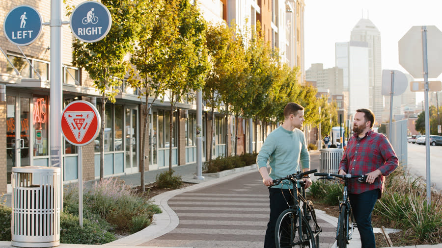 Two people standing next to bikes along the Indianapolis Cultural Trail; the city skyline is in the background.