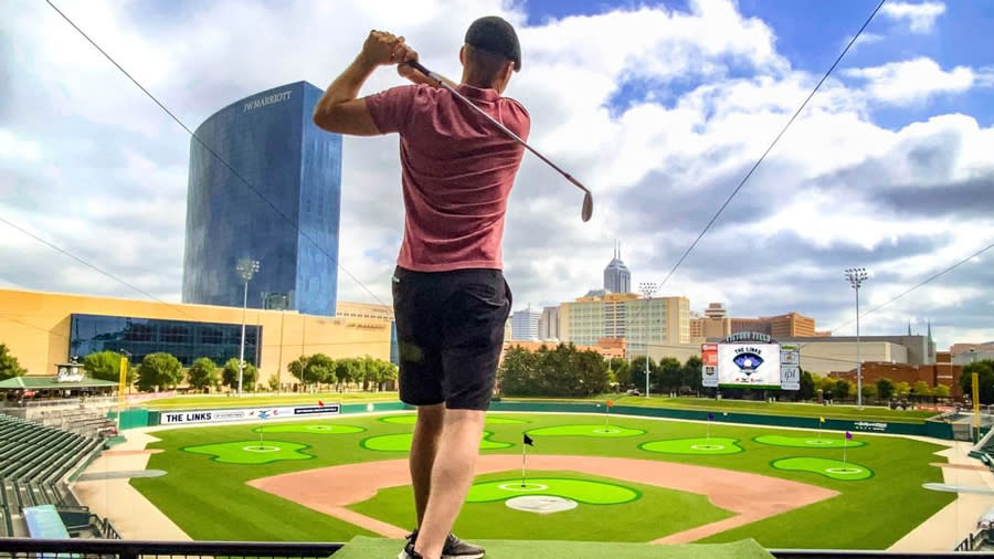 A man playing golf at Victory Field baseball field, with the Indy skyline in the background