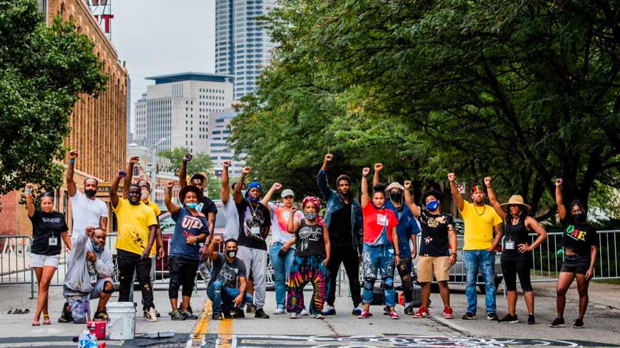 A group of artists stand over a #BlackLivesMatter mural. Their hands are held in the air in fists.