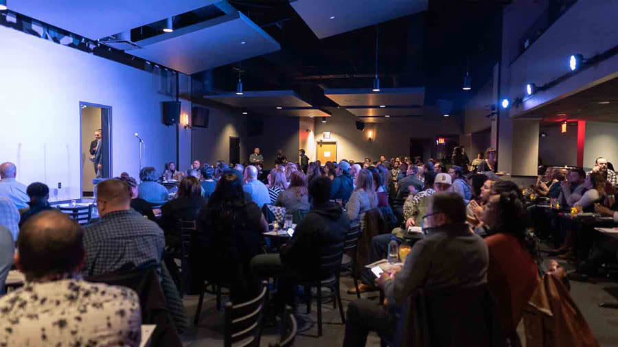 A group of people sit at chairs and tables at Helium Comedy Club. There is a performer on stage whom they are watching. 