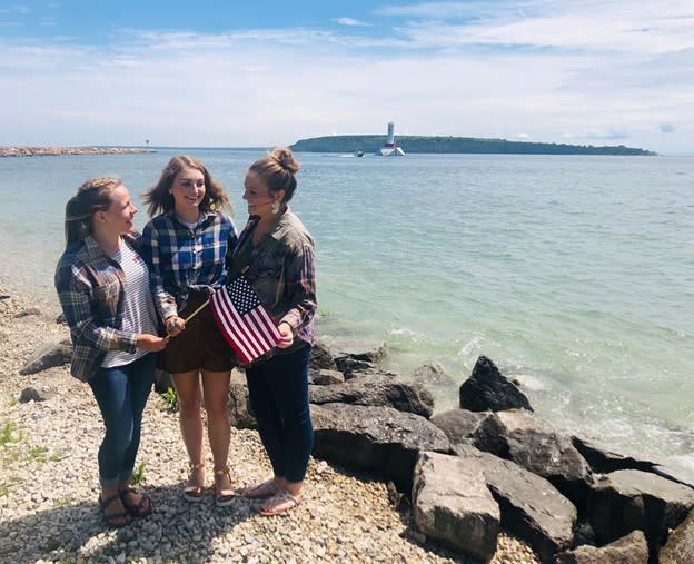 Three young women hold a small, American flag while standing along the water on a rocky Mackinac Island beach