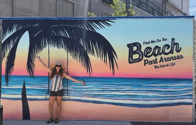 A woman stands, arms outstretched, in front of a mural that features a beach, a palm tree, and a swing. Text on the mural reads "Find Me On The Beach Port Aransas The Island Life"