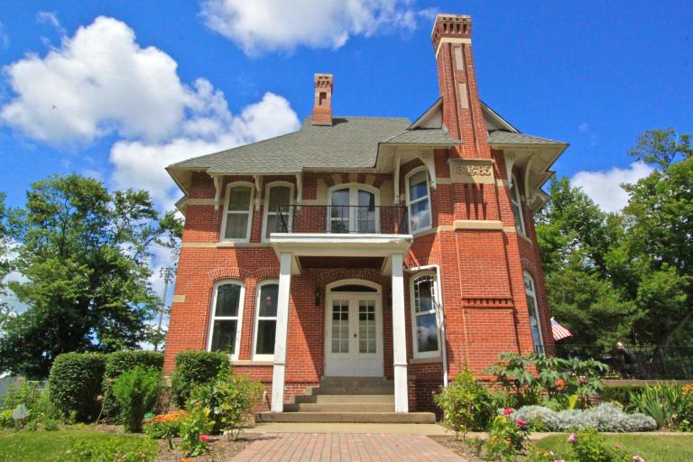 A red brick historic building with white columns. Trees and landscaping are on each side. The bright blue sky has white clouds.