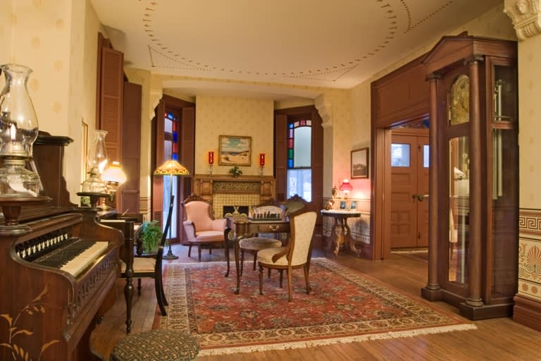 Interior of a historic building. A large carpet sits on wood floors. A piano, grandfather clock, fireplace, and dining room table sit in the room.