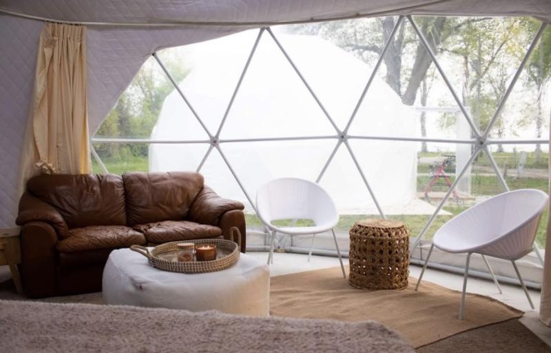 Inside a cozy geodesic dome, a brown leather couch, white chairs, and soft bedding sit in front of a large window overlooking trees and another dome outside.