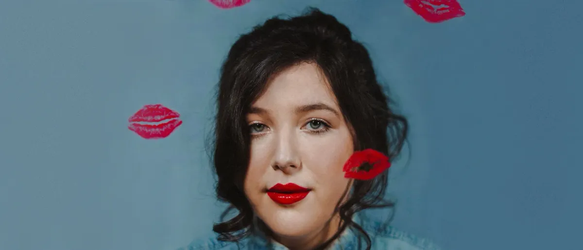 Lucy Dacus looks at the camera with a soft gaze, she is on a blue background with red lipstick kisses dotting the frame.