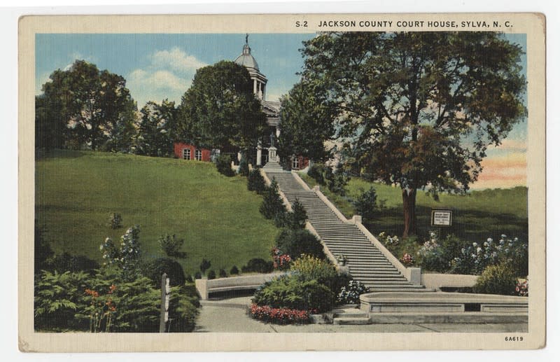 A vintage, colorized postcard depicting the Jackson County Court House in Sylva, N.C. The grand white building with a domed cupola sits atop a steep, lush green hill. A long, straight flight of concrete stairs leads directly up the hill to the entrance, flanked by manicured trees, shrubs, and flower beds.