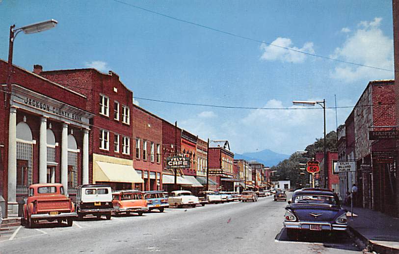 A vintage color postcard showing a bustling downtown street scene in Sylva, filled with parked cars from the 1960s. Brick storefronts line both sides of the street, featuring prominent signage for businesses like the columned "Jackson County Bank," "Velt's Cafe," and a "Phillips 66" gas station. Mountains fade into the horizon under a bright blue sky.