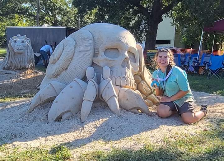 A woman kneels next to her sand sculpture, which looks like a human skull with snail shells instead of ears and crab legs on the bottom.