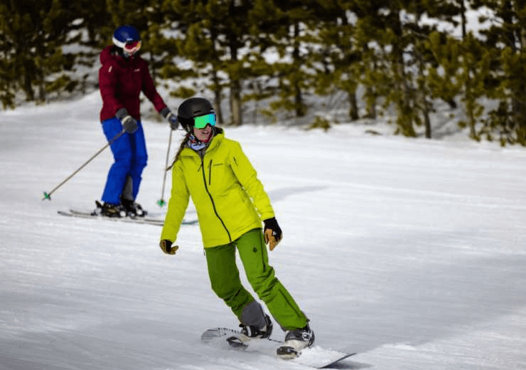 A snowboarder in lime-green attire gracefully carves through the snow, trailed by a skier in vibrant red and blue, both set against a serene landscape of snow-laden trees.