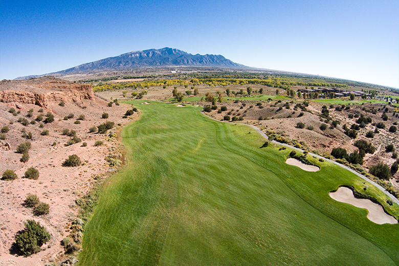 Aerial view of a golf course with lush greenery and sand traps, set against a backdrop of a large mountain and clear skies.