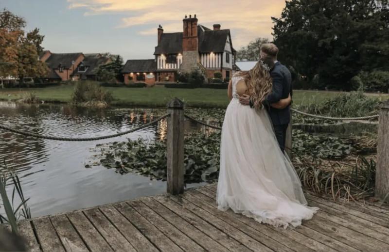 A bride and groom stand on a jetty, looking back over the lake at the beautiful Moat House Acton Trussell, Staffordshire