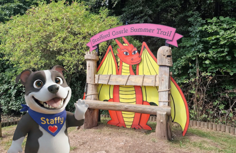 Staffy promoting the Stafford Castle Summer Trail next to a dragon-themed activity display at Stafford Castle.