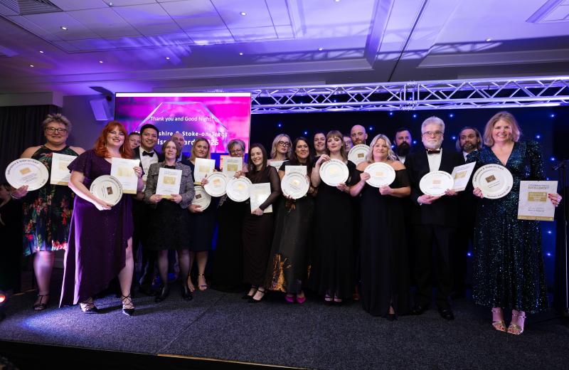 A group shot of the winners of the 2025 Staffordshire & Stoke-on-Trent Tourism Awards. There are about 20 people on a stage, wearing glamorous dresses and tuxedos, and holding trophies and certificates.
