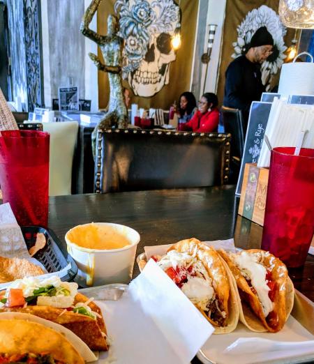 A table full of tacos, queso, red glasses, with funky art on the walls in the background at Agave and Rye in Covington, Ky.