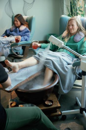 Two women lean back in reclining chairs, enjoying a relaxing foot soak. Both are dressed in robes with soft pillows and drinks in their hands.